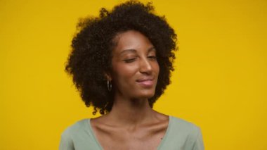 Doğal kıvırcık saçlı genç bir kadının portresi, neşeli bir şekilde göz kırpıyor ve parlak sarı bir arka plana gülümsüyor.