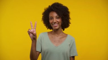 Kıvırcık saçlı genç bir kadının portresi. Elleriyle barış işareti yaparken sıcacık bir şekilde gülümsüyor.