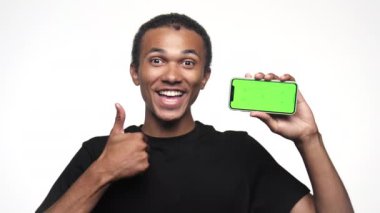 Siyah tişörtlü mutlu genç adam yeşil ekranlı akıllı telefonu gösteriyor ve beyaz arka planda başparmak işareti yapıyor.