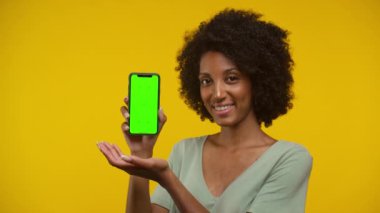Gülümseyen bir kadın elinde yeşil ekranlı bir akıllı telefon tutuyor. Özelleştirmeye hazır, sarı bir arka plana karşı.