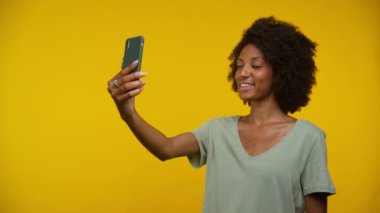 Bir kadın akıllı telefonuyla neşeli bir selfie çekiyor parlak sarı bir arka plana karşı, gülümsüyor ve poz veriyor.