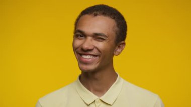 Neşeli bir genç adam ışıl ışıl sarı bir arka plana karşı durarak içten bir şekilde göz kırpıyor ve gülümsüyor.