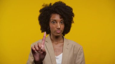 Bej rengi ceketli doğal kıvırcık saçlı kadın, uyarı jestinde yukarı doğru ciddi bir ifadeyle, sarı arka planda.