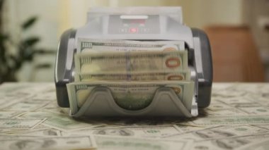 Para sayma makinesi 100 Amerikan banknotunu işliyor. Dolly zoom, ek para masanın etrafına saçılmış durumda..