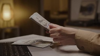 100 dolarlık banknotları açık bir dizüstü bilgisayarın üzerinde tutan ve sayan kişi, mali yönetim ve zenginliği sembolize ediyor.
