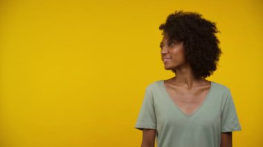 Yan tarafa bakarken gülümseyen genç bir kadın, parlak sarı bir arka planın önünde duruyor.