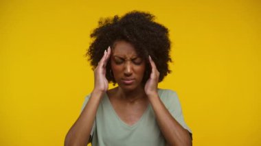Kıvırcık saçlı kadın iki eliyle başını tutuyor. Rahatsızlık ya da stres belirtileri gösteriyor. Parlak sarı bir arka planda duruyor.