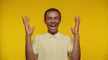 Neşeli genç bir adam ellerini kaldırıyor ve sarı bir arka planda sarı bir gömlek giyip heyecanını dile getiriyor.