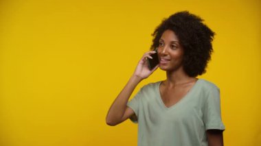 Kıvırcık saçlı bir kadın akıllı bir telefonla konuşuyor, gülümsüyor, parlak sarı bir arka plana karşı..