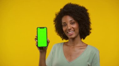 Kıvırcık saçlı gülümseyen kadın yeşil ekran maketli akıllı bir telefon tutuyor, parlak sarı bir arka planda..