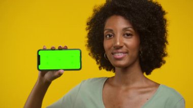 Kıvırcık saçlı bir kadın, yeşil ekran maketi olan akıllı bir telefonu sarı arka planda tutuyor..