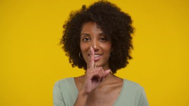 Kadın dudakları üzerinde parmak ile sessiz bir jest yapıyor, parlak sarı arka plana karşı sessizliği sembolize ediyor