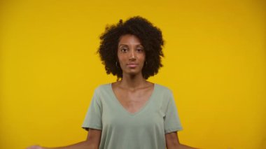 Doğal kıvırcık saçlı ve günlük kıyafetleri olan genç bir kadın umursamaz bir tavırla parlak sarı bir arka plana karşı omuz silkiyor.