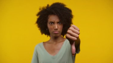Doğal kıvırcık saçlı genç bir kadın, parlak sarı bir arka plana karşı, onaylamadığını belirten, baş parmağını aşağı gösteren bir jest yapıyor.