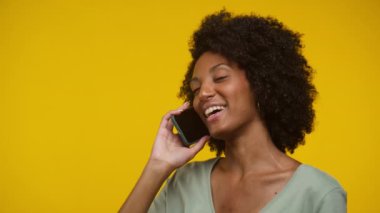Doğal kıvırcık saçlı genç bir kadın kulağına akıllı telefon tutarken gülümsüyor, sarı arka planda hoş bir sohbet ediyor.