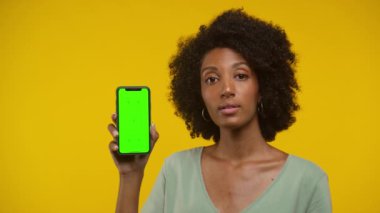 Doğal kıvırcık saçlı, elinde yeşil ekranlı akıllı bir telefon tutan genç bir kadın, sarı bir arka plana karşı şüphecilik gösteriyor.