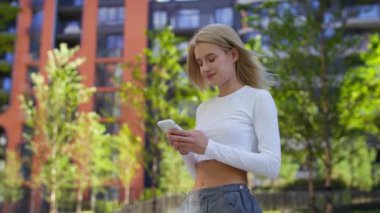 Sarışın bir kadın açık havada akıllı telefonlara bakıyor. Bulanık apartmanlı kentsel ortam.