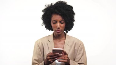 Bej ceketli genç bir kadın, beyaz arka planda izole edilmiş, hoşnutsuz bir ifadeyle akıllı telefonuna bakıyor.