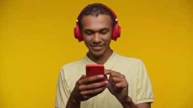 Kırmızı kulaklık takan ve akıllı telefonuna bakarken şaşırmış bir ifadeyle tepki veren genç bir adam, parlak sarı bir arka plana karşı duruyor.