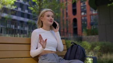 Beyaz tişörtlü genç bir kadın, rahatlamış bir ifadeyle parkta bankta otururken telefonla konuşuyor..