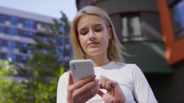 Beyaz bluzlu genç bir kadın açık havada şehir ortamında akıllı telefonunu kullanmaya odaklanmış.