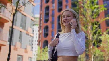 Beyaz bir bluz giyen gülümseyen genç bir kadın modern binalarla dolu bir şehirde yürürken akıllı telefonuyla konuşuyor..