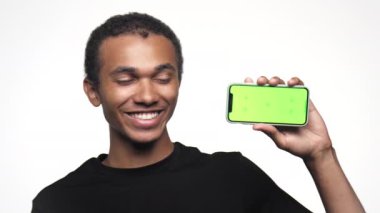 Siyah tişörtlü mutlu genç adam yeşil ekranlı akıllı bir telefon gösteriyor, beyaz arka plana karşı baş parmak işareti yapıyor.