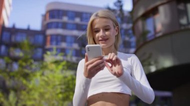 Endişeli kadın akıllı telefon ekranına bakıyor, arka planda binalar ve yeşillik olan bir şehir bölgesinde açık havada..
