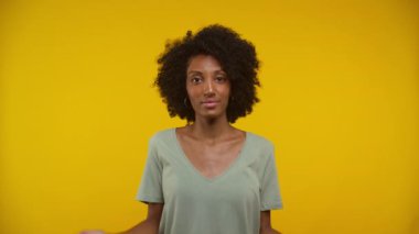 Yeşil gömlekli bir kadın, parlak sarı bir arkaplan karşısında şaşkın bir ifadeyle omuz silkiyor..