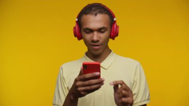 Kırmızı kulaklık takan genç adam, canlı sarı bir arka plana karşı akıllı telefon kullanırken gülümsüyor..