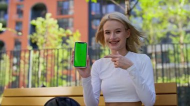Gülümseyen kadın şehir ortamında yeşil ekranlı akıllı telefon gösteriyor..