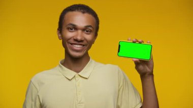 Adam yeşil ekranlı akıllı telefonu tutarken gülümsüyor ve sarı arka planda baş parmak işareti yapıyor..