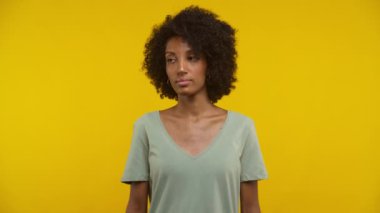 Doğal kıvırcık saçlı ve halka küpeli genç bir kadının yakın plan fotoğrafı. Sağlam sarı bir arka plana karşı güvenle ayakta duruyor.