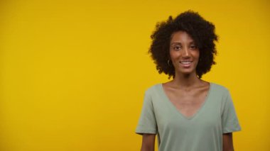 Doğal kıvırcık saçlı ve halka küpeli neşeli genç bir kadının portresi. Kopya alanına bakıyor ve parlak sarı bir arka plana karşı onayıyla başını sallıyor.