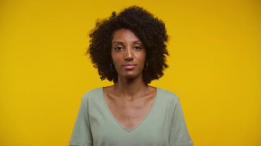 Doğal kıvırcık saçlı ve halkalı küpeli genç bir kadın, bilgi ve seçimin eksikliğini sembolize etmek için omuzlarını silkiyor, canlı bir sarıya karşı duruyor.
