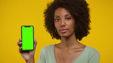 Parlak sarı bir arkaplan karşısında duran, yeşil ekranlı akıllı telefonu tutan ve gösteren bir kadın.