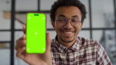 Gözlüklü ve ekose gömlekli neşeli genç adam, kameraya yakın yeşil ekranlı akıllı bir telefonu tutuyor, modern bir iç mekan ortamında..