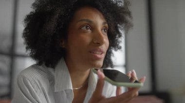 Doğal kıvırcık saçlı profesyonel bir kadın, düğmeli gömlek giyiyor, akıllı telefonunu ağzına yakın tutuyor ve modern ofis ortamında sesli mesaj kaydediyor.