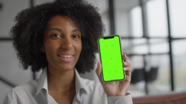 Doğal kıvırcık saçlı, çizgili gömlek giyen, elinde yeşil ekranlı akıllı bir telefonla gülümseyen ve modern ofis ortamında sunum yapan genç bir kadın..