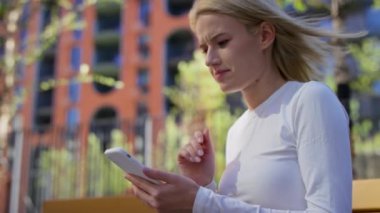 Beyaz bluzlu sarışın bir kadın akıllı telefonuyla internette geziniyor, modern bir şehir parkında bankta oturuyor..