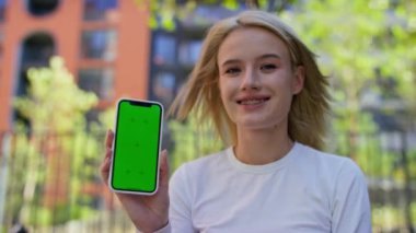 Telleri olan sarışın genç bir kadın yeşil ekranlı akıllı bir telefonu tutuyor, modern bir açık hava parkı ortamında gülümsüyor..