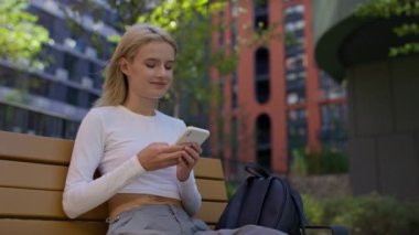 Beyaz bluzlu sarışın bir kadın akıllı telefonuyla yazarken kafası karışık bir ifadeyle modern bir şehir parkında oturuyor..