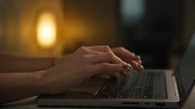 Arka planda sıcak ortam ışığı olan rahat bir ortamda dizüstü bilgisayarda yazı yazan ellerin yakın çekimi.