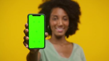 Bir kadın yeşil ekran odaklı akıllı bir telefon tutuyor, arka planda sarı bir fona gülümsüyor..