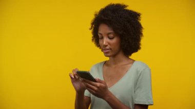 Bir kadın, parlak sarı bir arkaplanda dikilirken akıllı telefonuyla iletişim kurar, odaklanmış görünür..