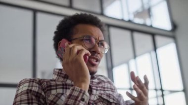 Gözlüklü bir adam telefonla konuşurken hayal kırıklığını ifade ediyor, modern bir ofis ortamında hareketler yapıyor..