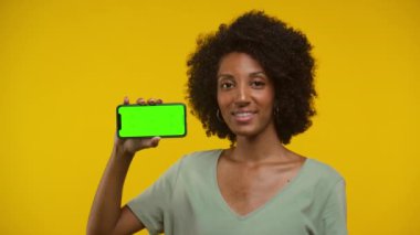 Elinde yeşil ekranlı akıllı telefon tutan gülümseyen genç bir kadın ve sarı arka plana karşı onaylı başıyla başını sallıyor..