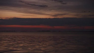 Sakin suları ve ufuktaki dramatik bulutlar tarafından çerçevelenmiş canlı turuncu bir gökyüzünü gösteren sakin bir alacakaranlık sahnesi..