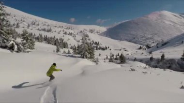 Yalnız bir snowboardcu açık mavi gökyüzünün altında kar kaplı bir dağ yamacında taze tozu oyuyor..