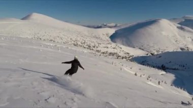 Yalnız bir snowboardcu uçsuz bucaksız, kar kaplı bir dağda, açık mavi bir gökyüzünün altında pürüzsüz bir yol kazıyor..